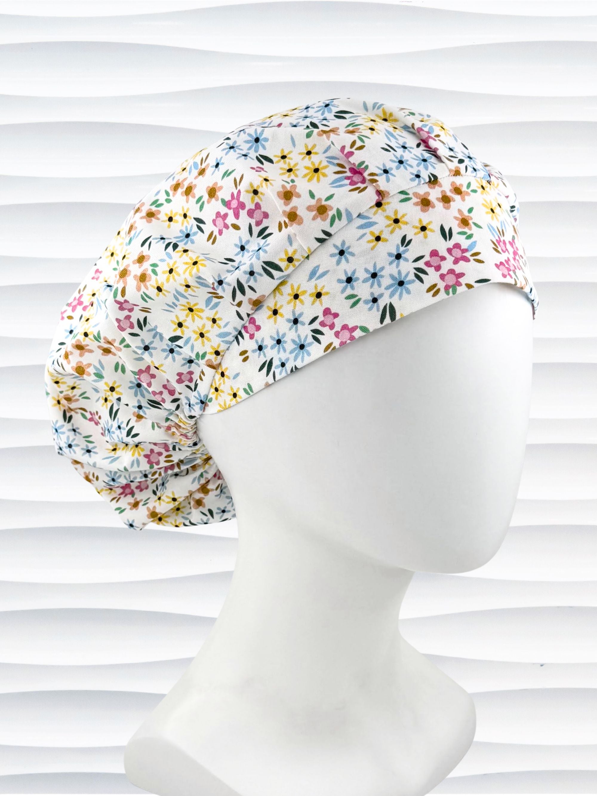 A white fabric bouffant style surgical cap by JensCaps with a colorful floral print, displayed on a white mannequin head against a white wavy background.