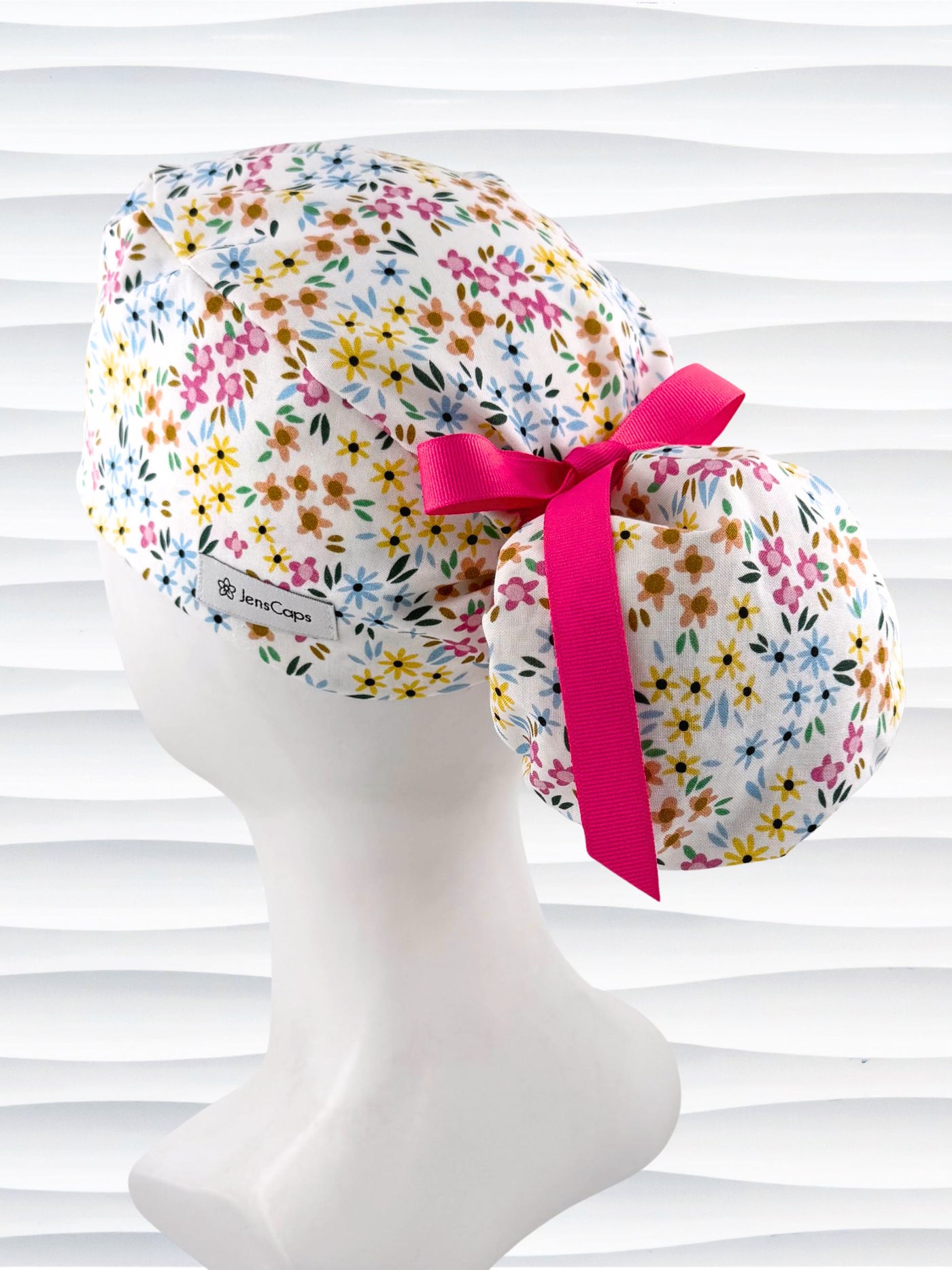 Ponytail style surgical scrub cap by JensCaps with small flowers in pinks, blues, and yellow on white cotton fabric, tied with a grosgrain ribbon, and displayed on a white mannequin head.