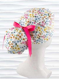 Ponytail style surgical scrub cap by JensCaps with small flowers in pinks, blues, and yellow on white cotton fabric, tied with a grosgrain ribbon, and displayed on a white mannequin head.