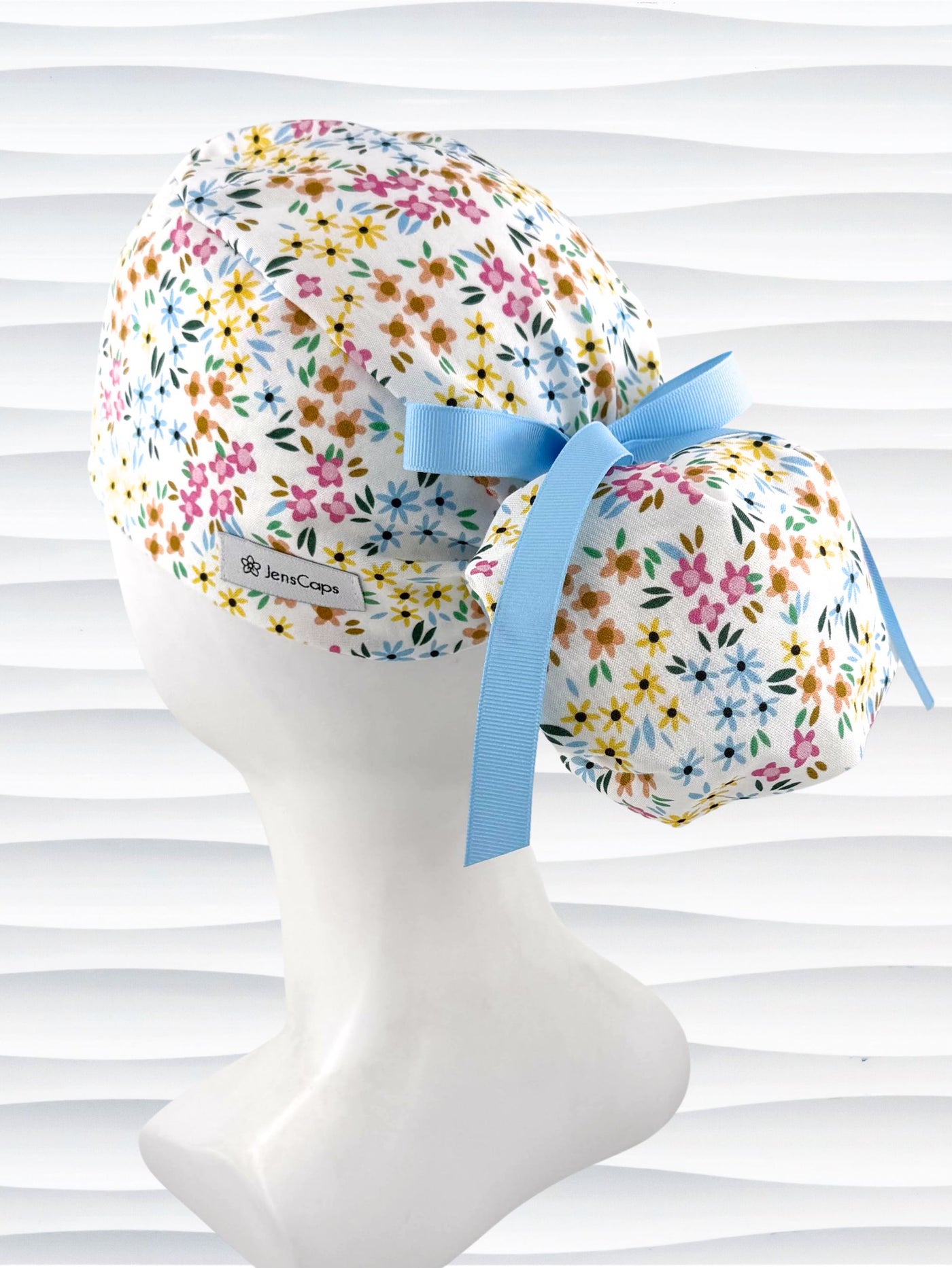 Ponytail style surgical scrub cap by JensCaps with small flowers in pinks, blues, and yellow on white cotton fabric, tied with a grosgrain ribbon, and displayed on a white mannequin head.