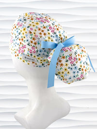 Ponytail style surgical scrub cap by JensCaps with small flowers in pinks, blues, and yellow on white cotton fabric, tied with a grosgrain ribbon, and displayed on a white mannequin head.