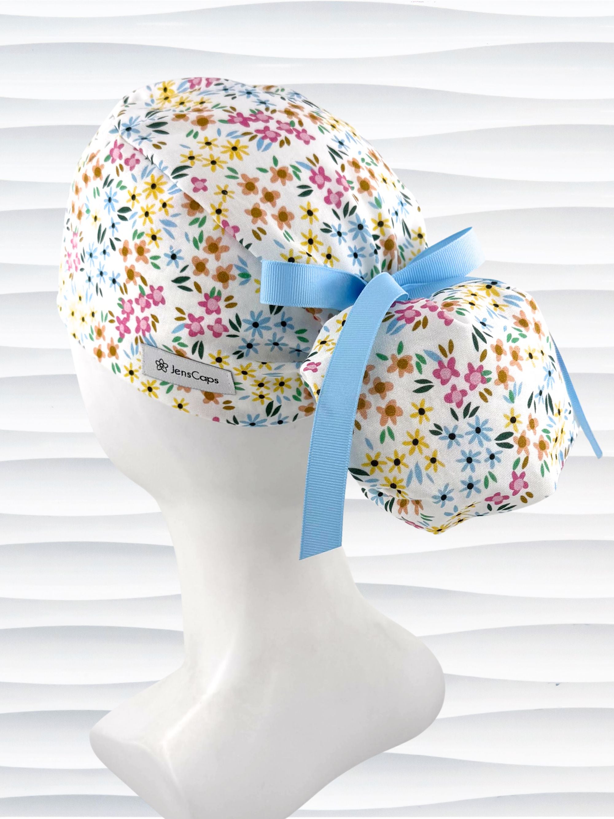 Ponytail style surgical scrub cap by JensCaps with small flowers in pinks, blues, and yellow on white cotton fabric, tied with a grosgrain ribbon, and displayed on a white mannequin head.