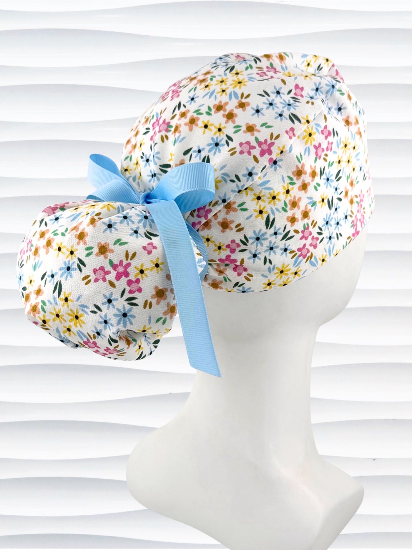 Ponytail style surgical scrub cap by JensCaps with small flowers in pinks, blues, and yellow on white cotton fabric, tied with a grosgrain ribbon, and displayed on a white mannequin head.