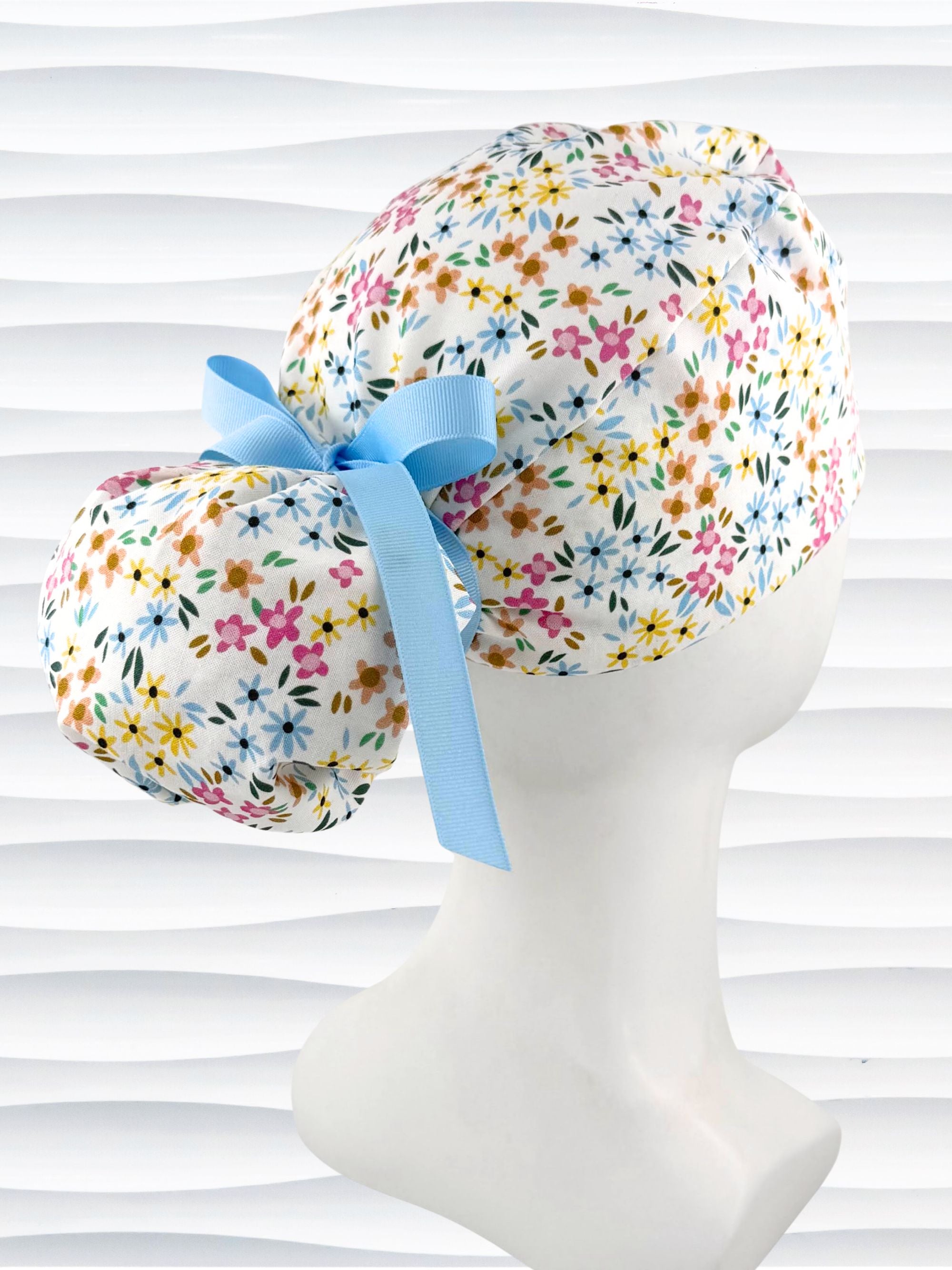 Ponytail style surgical scrub cap by JensCaps with small flowers in pinks, blues, and yellow on white cotton fabric, tied with a grosgrain ribbon, and displayed on a white mannequin head.
