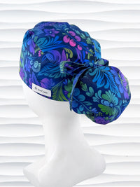 Ponytail style surgical scrub cap by JensCaps with an allover floral pattern in dark blue, purple, emerald, and turquoise, finished with a ribbon tie, and displayed on a mannequin head.