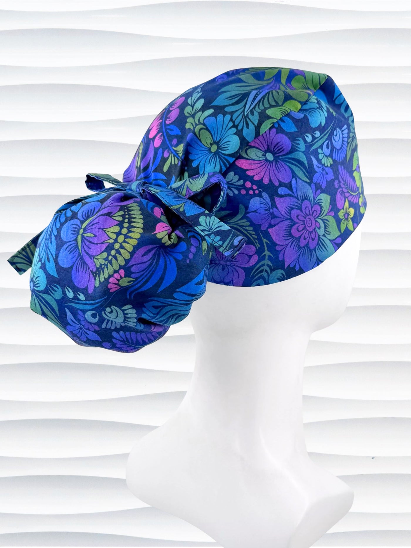 Ponytail style surgical scrub cap by JensCaps with an allover floral pattern in dark blue, purple, emerald, and turquoise, finished with a ribbon tie, and displayed on a mannequin head.