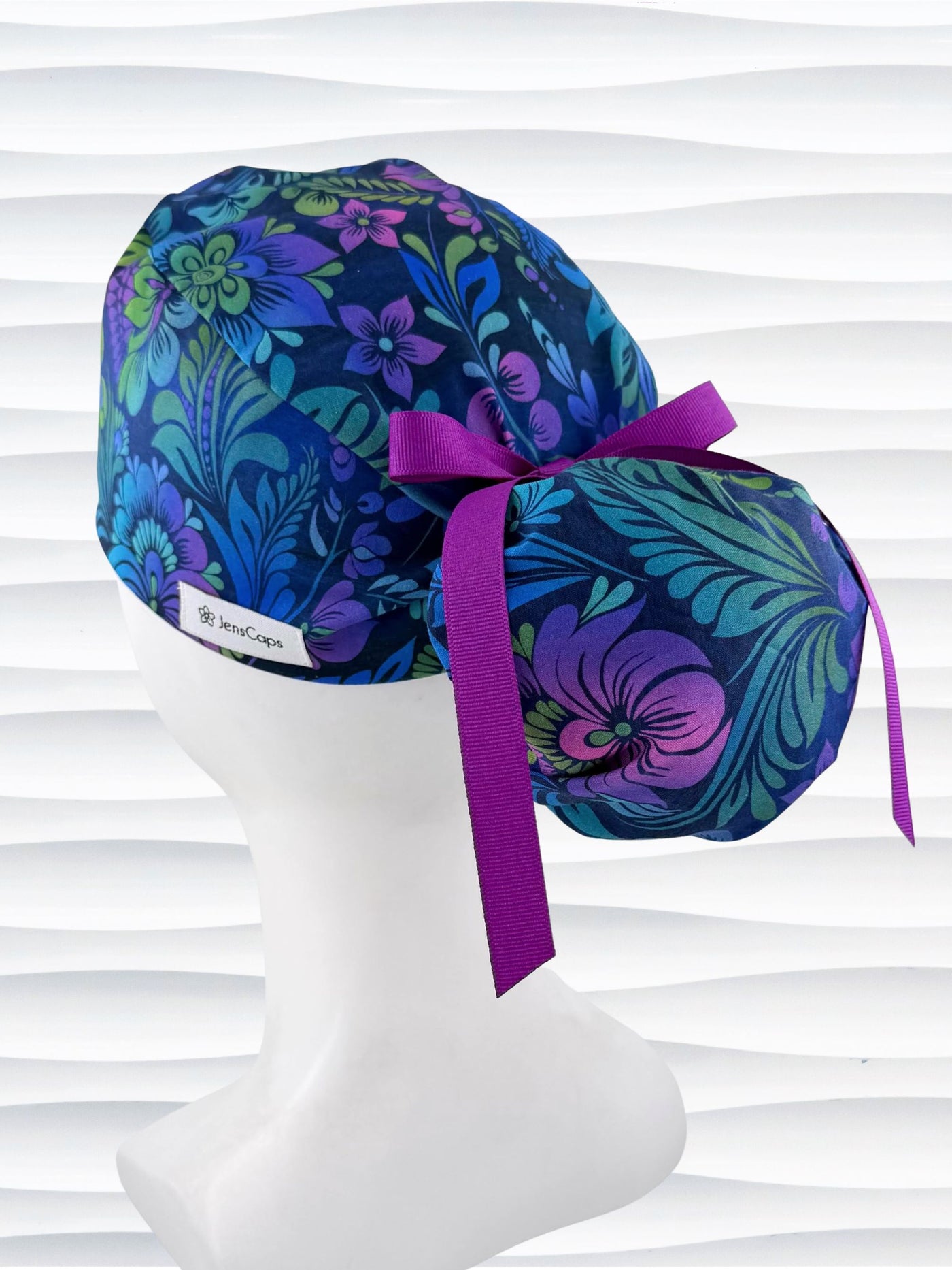 Ponytail style surgical scrub cap by JensCaps with an allover floral pattern in dark blue, purple, emerald, and turquoise, finished with a ribbon tie, and displayed on a mannequin head.