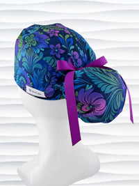 Ponytail style surgical scrub cap by JensCaps with an allover floral pattern in dark blue, purple, emerald, and turquoise, finished with a ribbon tie, and displayed on a mannequin head.