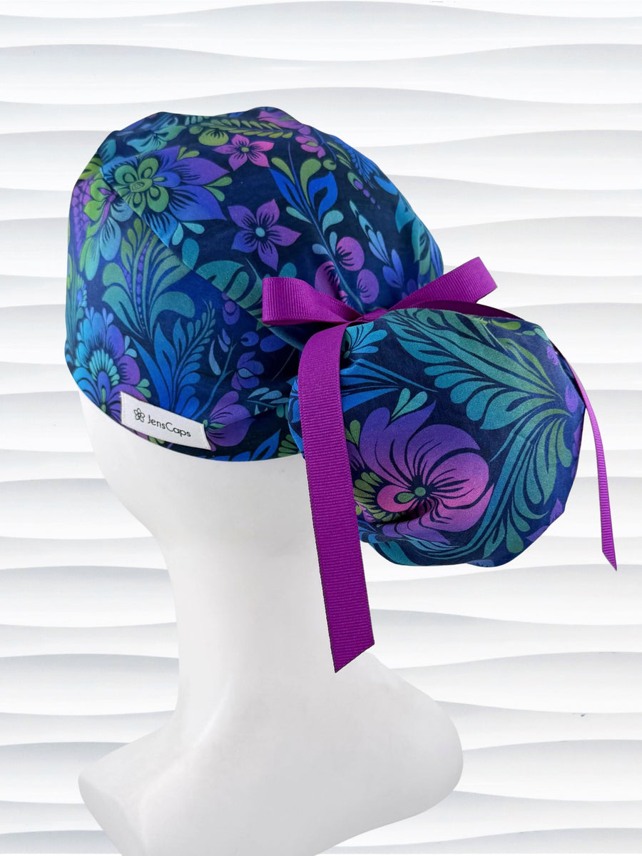 Ponytail style surgical scrub cap by JensCaps with an allover floral pattern in dark blue, purple, emerald, and turquoise, finished with a ribbon tie, and displayed on a mannequin head.