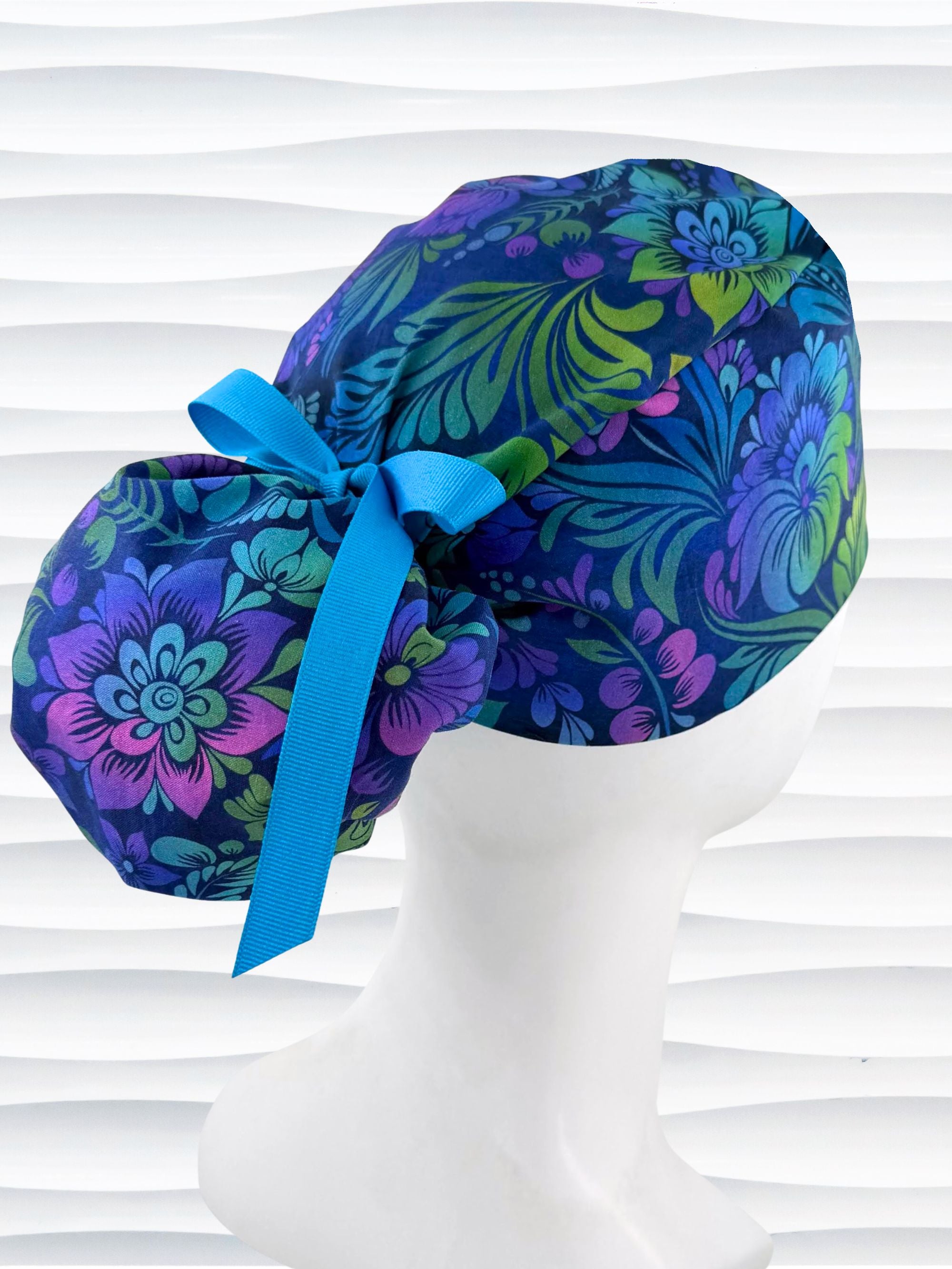 Ponytail style surgical scrub cap by JensCaps with an allover floral pattern in dark blue, purple, emerald, and turquoise, finished with a ribbon tie, and displayed on a mannequin head.