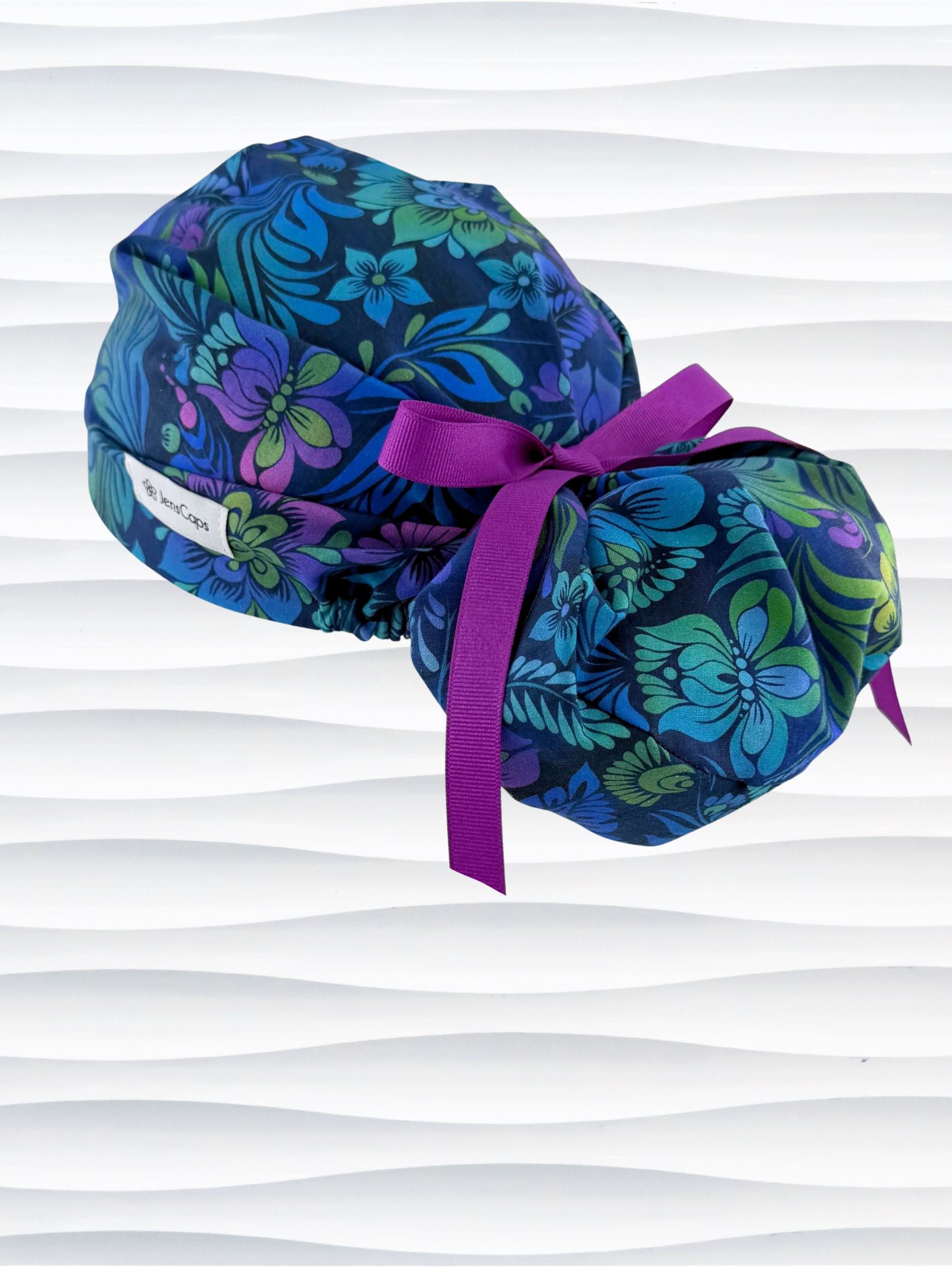 Euro Pony style surgical scrub cap by JensCaps with an allover floral pattern in dark blue, purple, emerald, and turquoise and displayed on a mannequin head.