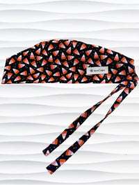A black tie back style surgical scrub cap with a pink, orange, and white candy corn pattern displayed on a white mannequin head. The brand name 'JensCaps' is visible on a white label sewn onto the side of the cap.