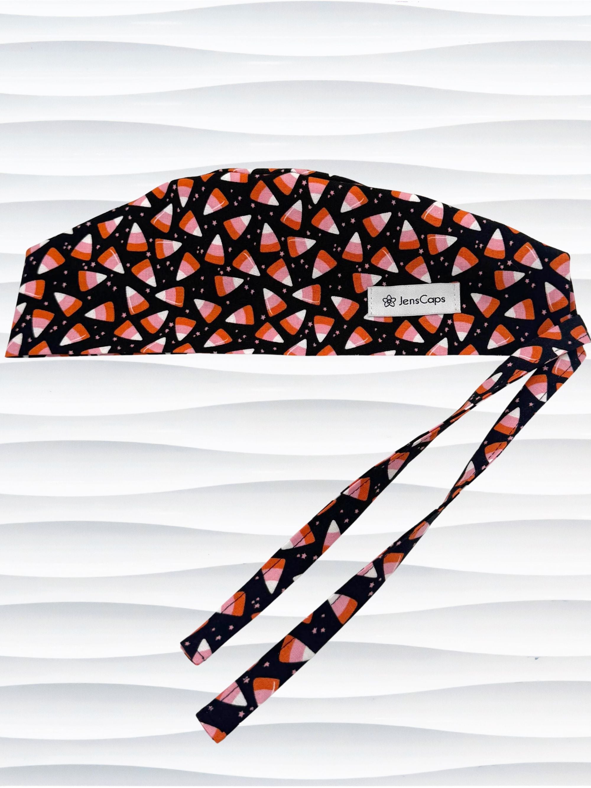 A black tie back style surgical scrub cap with a pink, orange, and white candy corn pattern displayed on a white mannequin head. The brand name 'JensCaps' is visible on a white label sewn onto the side of the cap.