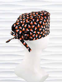 A black tie back style surgical scrub cap with a pink, orange, and white candy corn pattern displayed on a white mannequin head. The brand name 'JensCaps' is visible on a white label sewn onto the side of the cap.