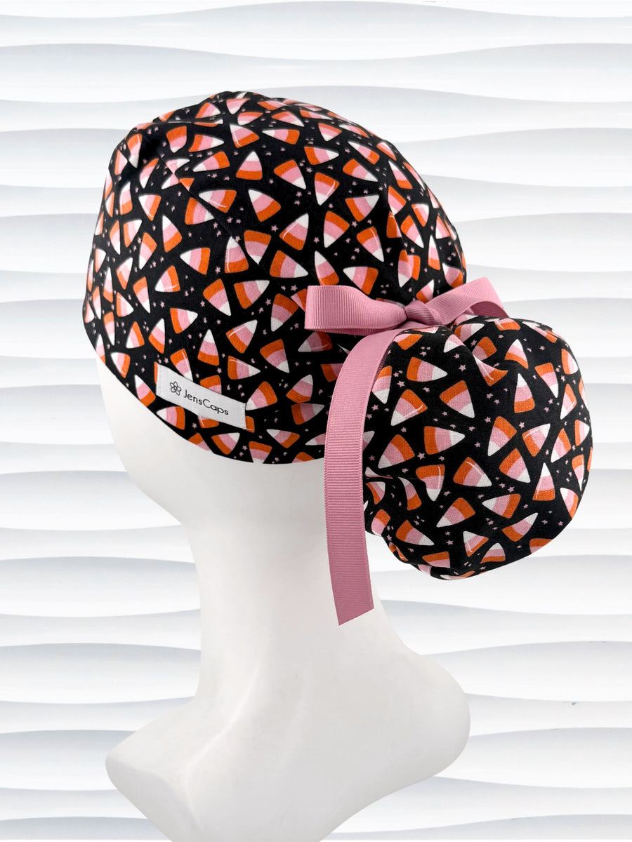A black ponytail style scrub cap by JensCaps with a candy corn pattern in pink, orange, and white, featuring a ribbon bow and displayed on a white mannequin head.