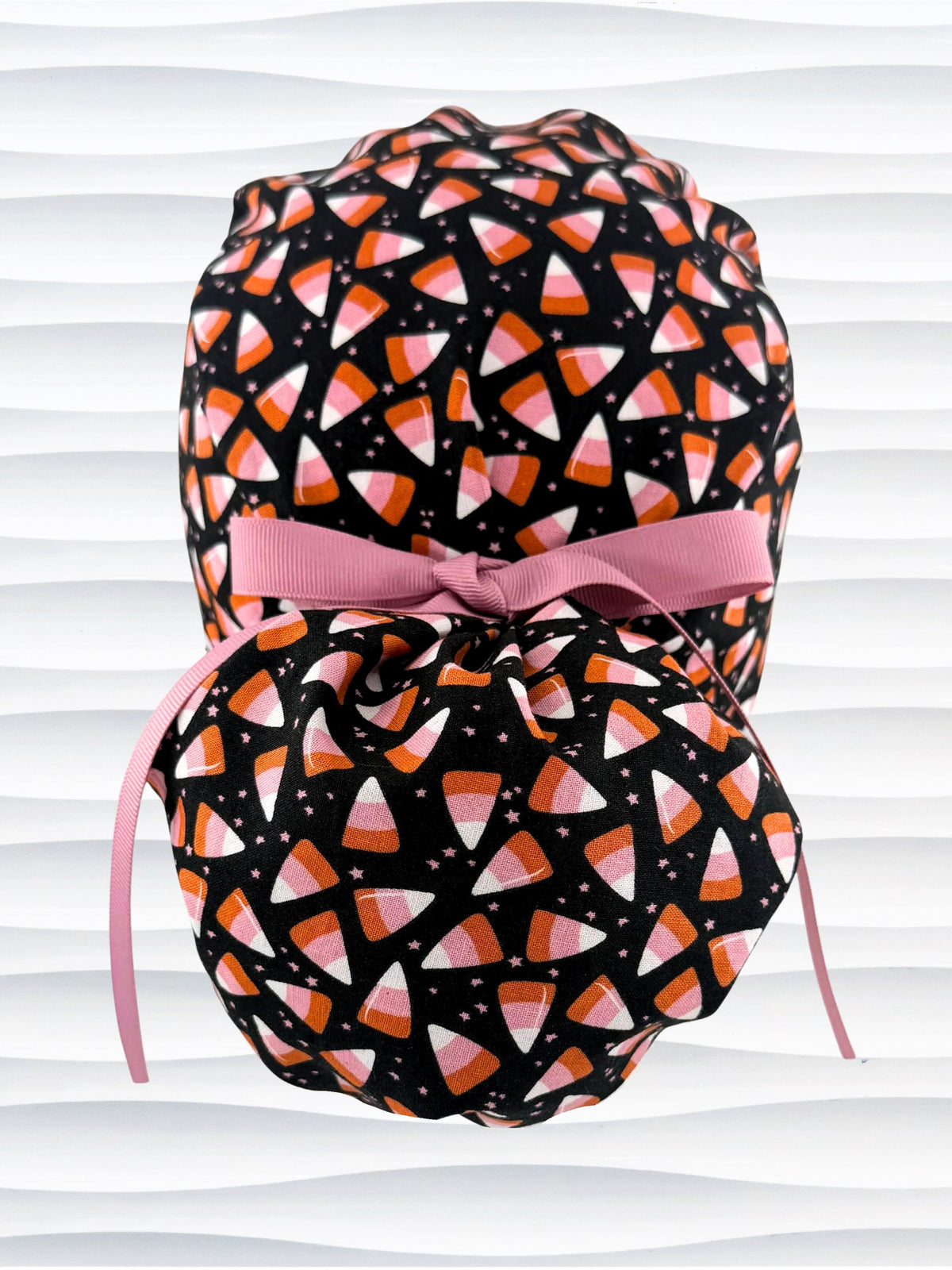 A black ponytail style scrub cap by JensCaps with a candy corn pattern in pink, orange, and white, featuring a ribbon bow and displayed on a white mannequin head.