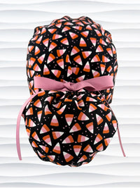 A black ponytail style scrub cap by JensCaps with a candy corn pattern in pink, orange, and white, featuring a ribbon bow and displayed on a white mannequin head.