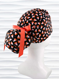 A black ponytail style scrub cap by JensCaps with a candy corn pattern in pink, orange, and white, featuring a ribbon bow and displayed on a white mannequin head.