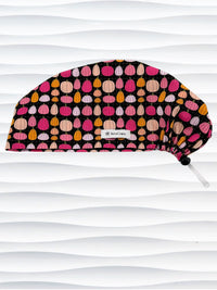 A black fabric Euro Pixie styles surgical scrub cap by JensCaps with a colorful pattern of pink, orange, and beige pumpkins displayed on a white mannequin head against a white wavy background.