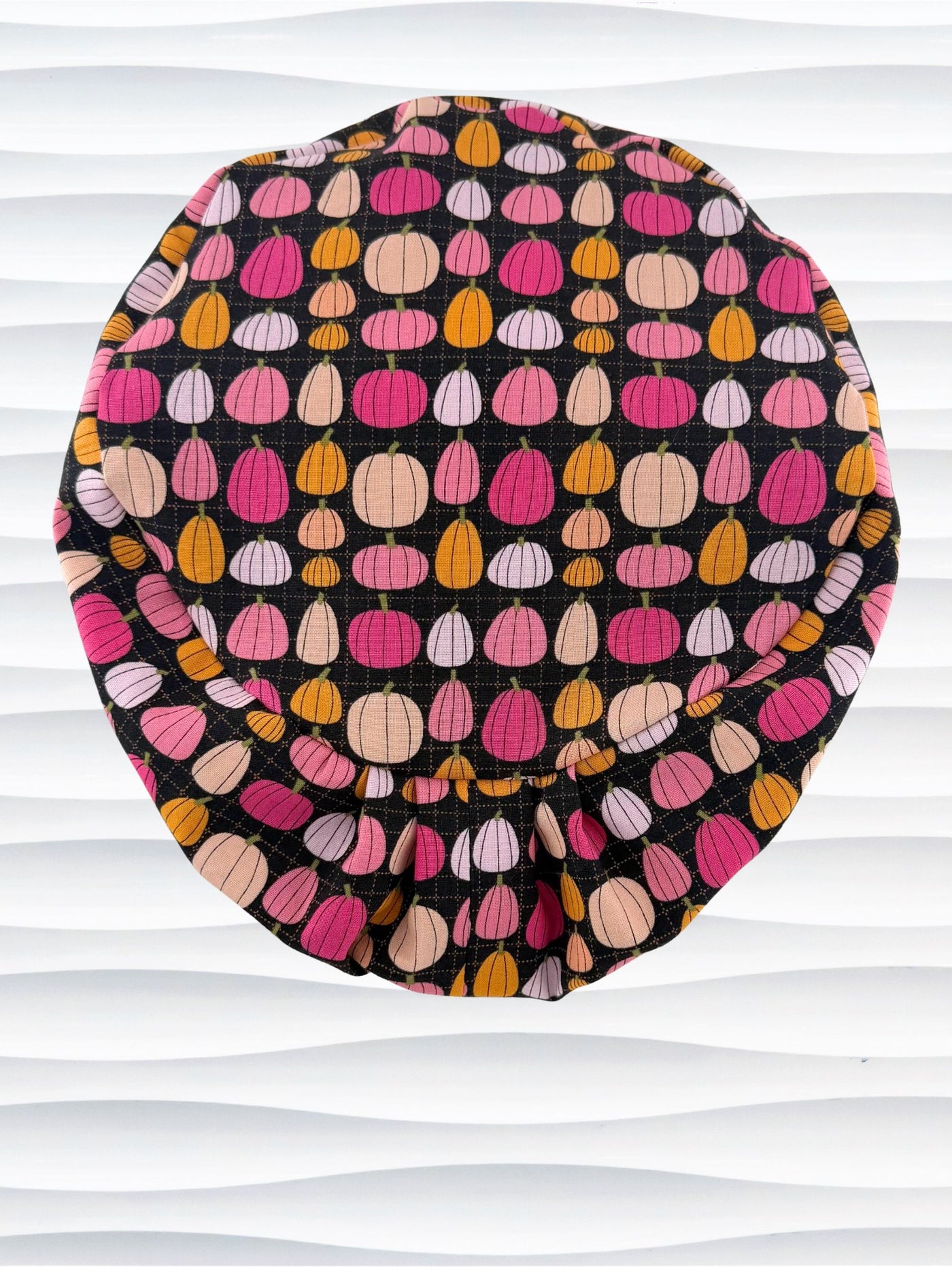 A black fabric Euro Pixie styles surgical scrub cap by JensCaps with a colorful pattern of pink, orange, and beige pumpkins displayed on a white mannequin head against a white wavy background.