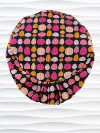 A black fabric Euro Pixie styles surgical scrub cap by JensCaps with a colorful pattern of pink, orange, and beige pumpkins displayed on a white mannequin head against a white wavy background.