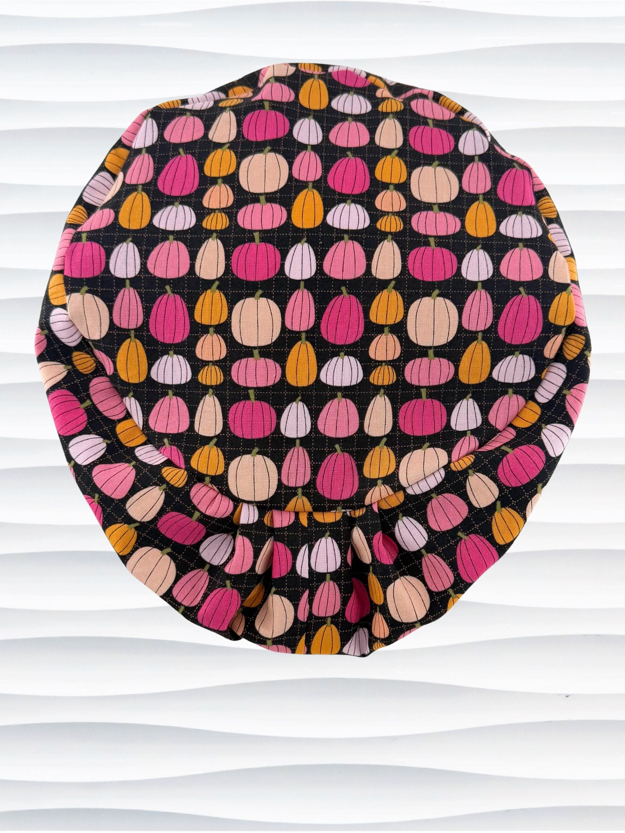 A black fabric Euro Pixie styles surgical scrub cap by JensCaps with a colorful pattern of pink, orange, and beige pumpkins displayed on a white mannequin head against a white wavy background.