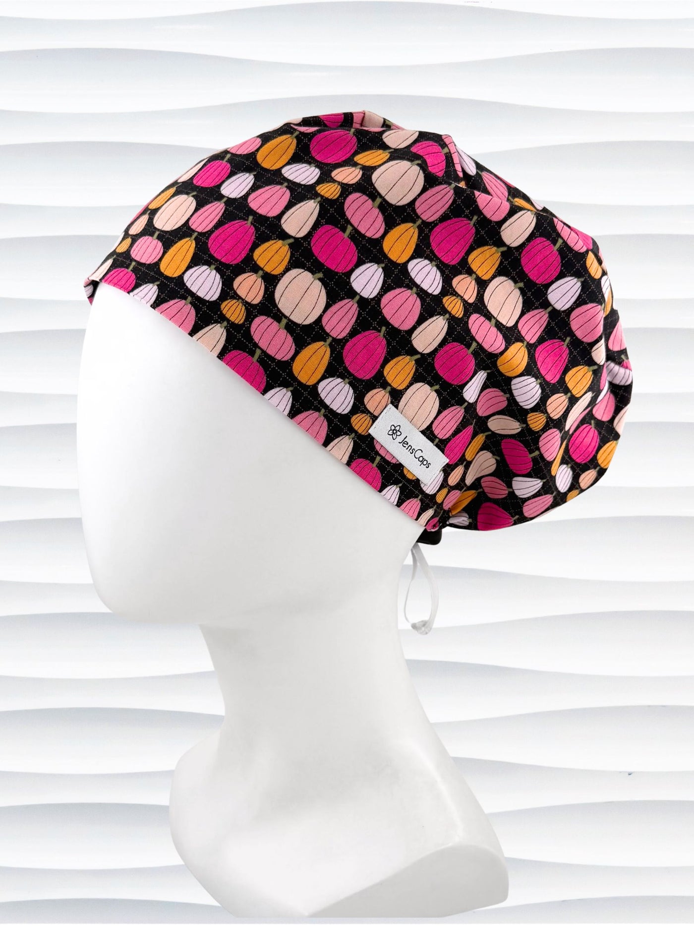 A black fabric Euro Pixie styles surgical scrub cap by JensCaps with a colorful pattern of pink, orange, and beige pumpkins displayed on a white mannequin head against a white wavy background.