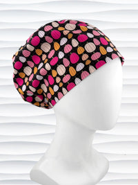 A black fabric Euro Pixie styles surgical scrub cap by JensCaps with a colorful pattern of pink, orange, and beige pumpkins displayed on a white mannequin head against a white wavy background.