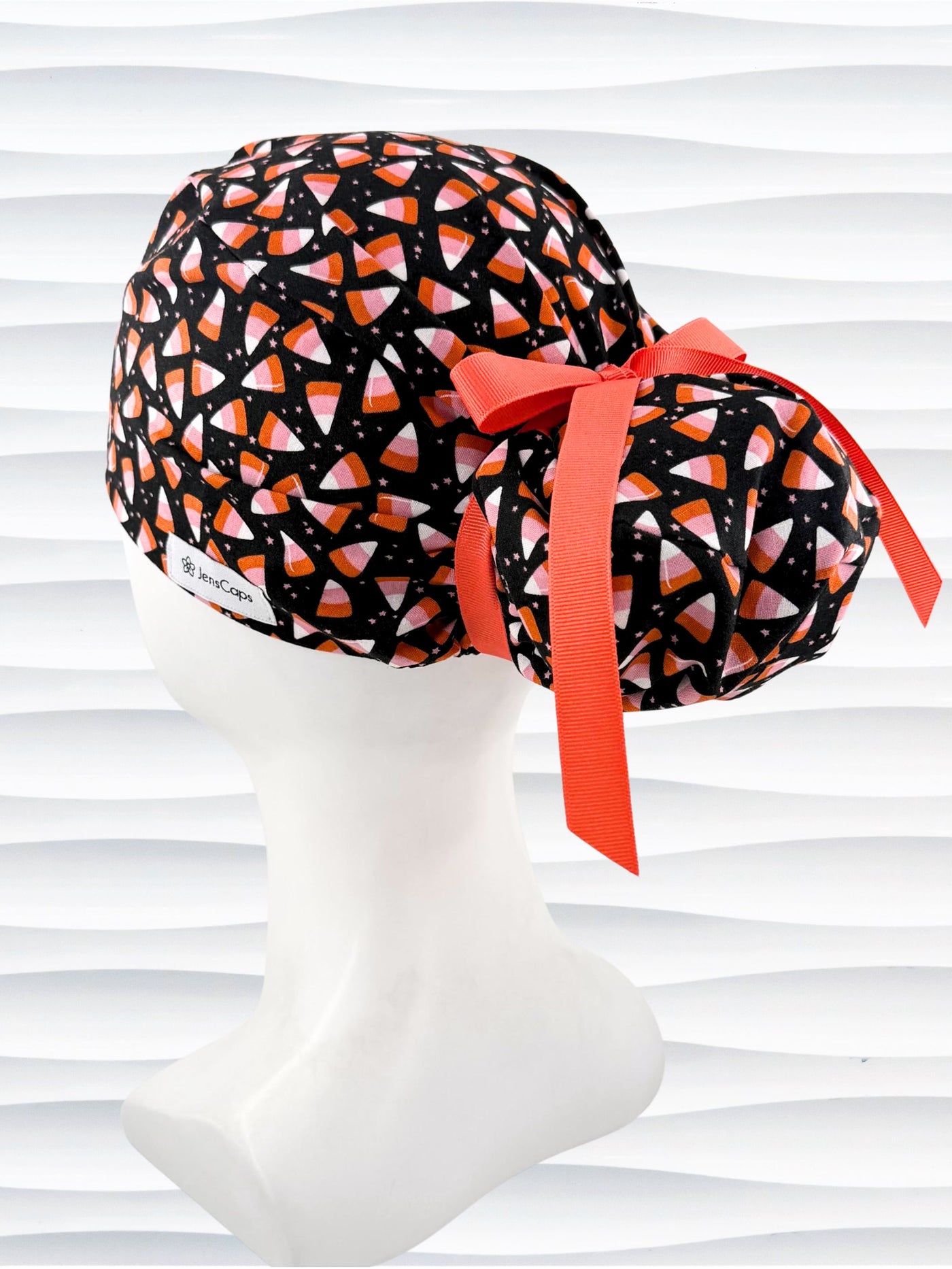 A black Ponytail style scrub cap by JensCaps with a pink, white, and orange  candy corn pattern, featuring a ribbon bow and displayed on a white mannequin head against a white wavy background.