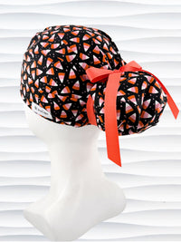 A black Ponytail style scrub cap by JensCaps with a pink, white, and orange  candy corn pattern, featuring a ribbon bow and displayed on a white mannequin head against a white wavy background.