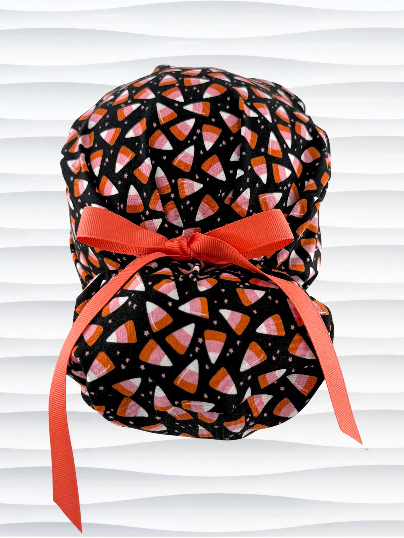 A black Ponytail style scrub cap by JensCaps with a pink, white, and orange  candy corn pattern, featuring a ribbon bow and displayed on a white mannequin head against a white wavy background.