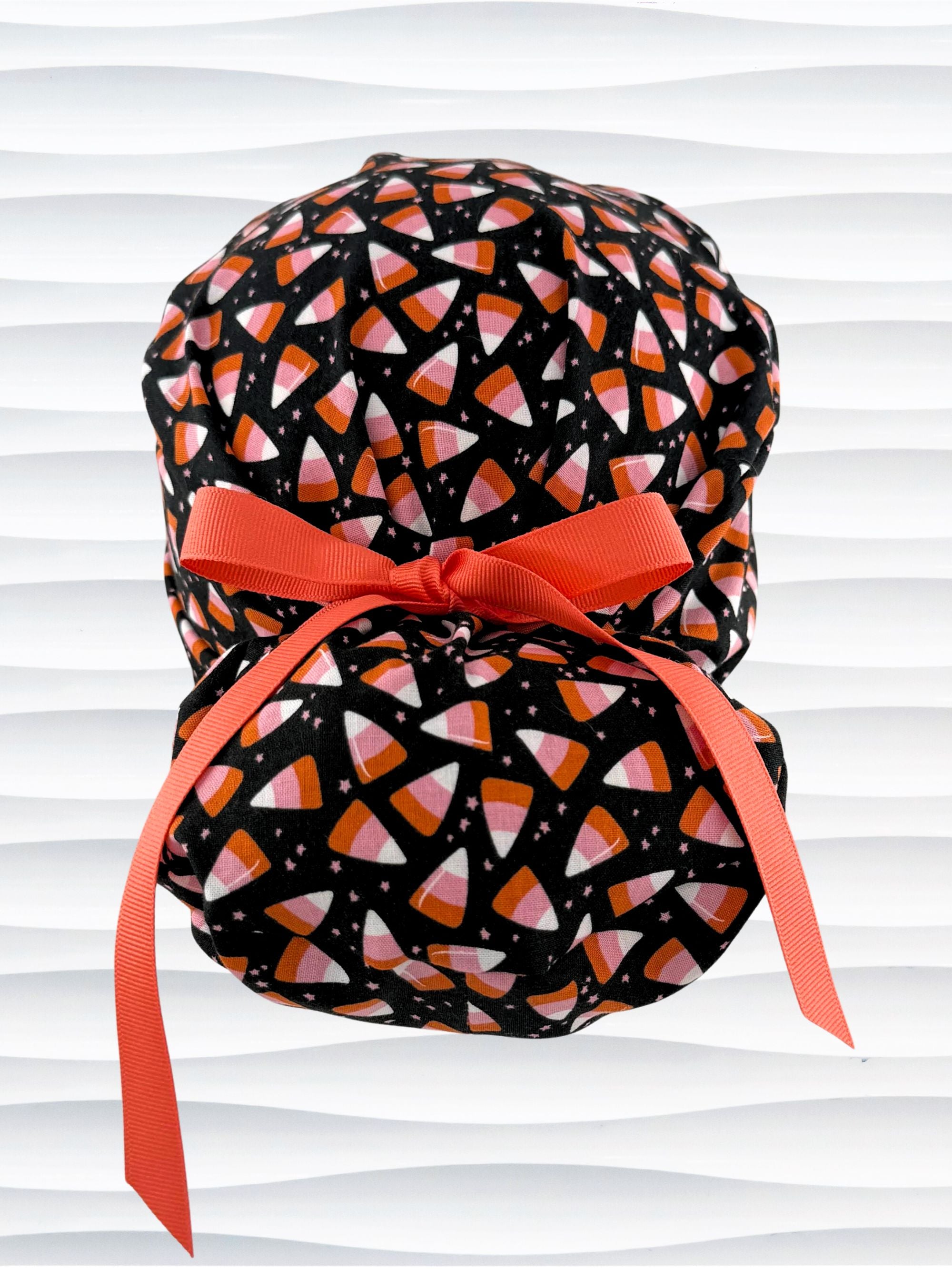 A black Ponytail style scrub cap by JensCaps with a pink, white, and orange  candy corn pattern, featuring a ribbon bow and displayed on a white mannequin head against a white wavy background.