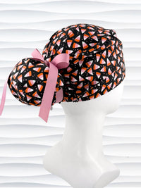 A black Ponytail style scrub cap by JensCaps with a pink, white, and orange  candy corn pattern, featuring a ribbon bow and displayed on a white mannequin head against a white wavy background.