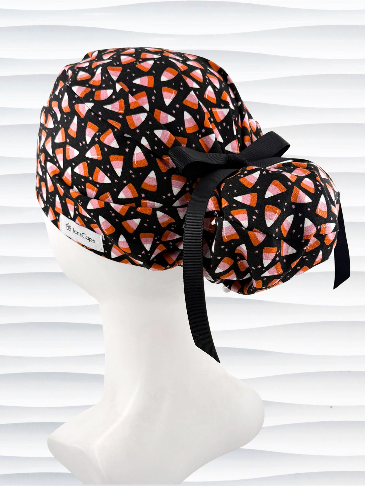 A black ponytail style scrub cap by JensCaps with a candy corn pattern in pink, orange, and white, featuring a ribbon bow and displayed on a white mannequin head.
