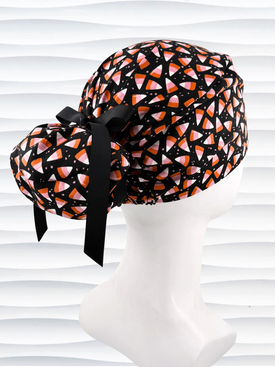 A black Ponytail style scrub cap by JensCaps with a pink, white, and orange  candy corn pattern, featuring a ribbon bow and displayed on a white mannequin head against a white wavy background.