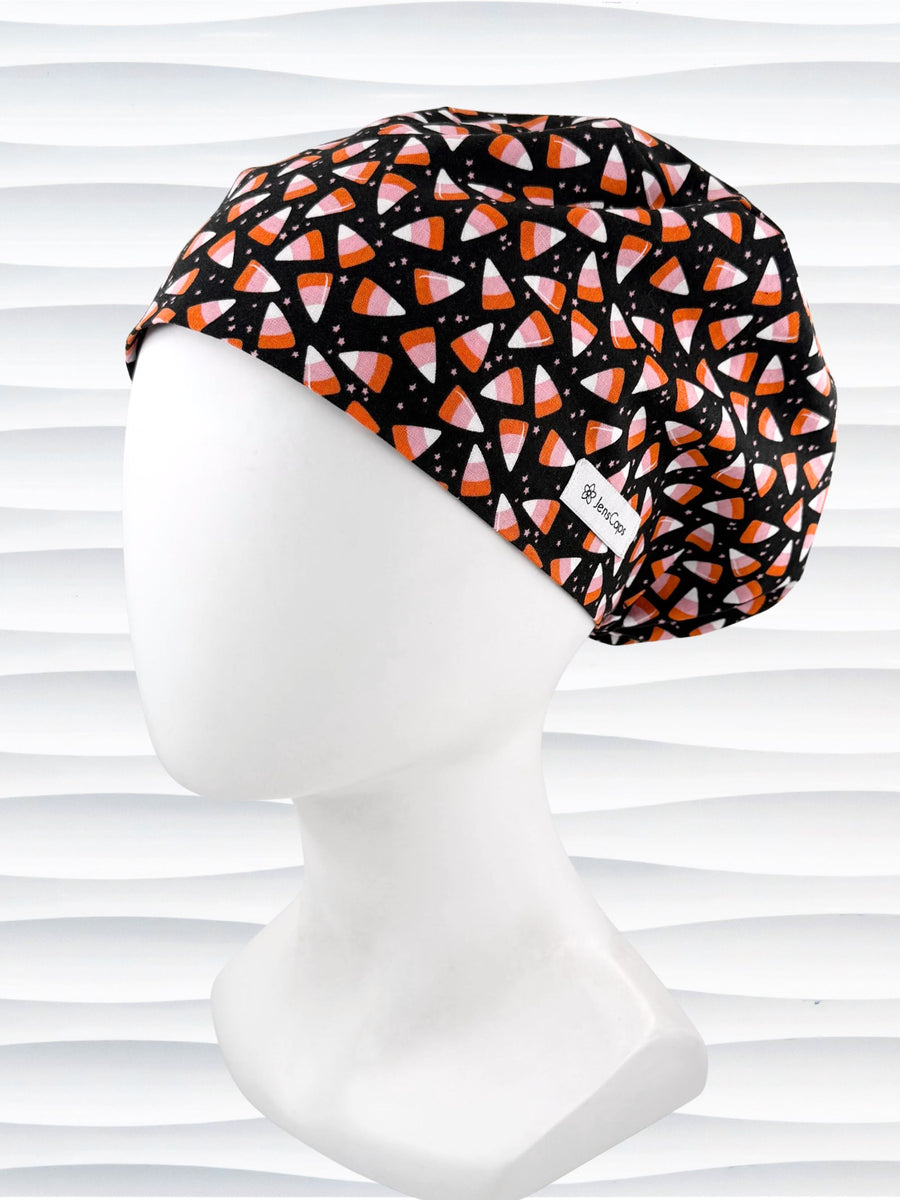 A Pixie style black scrub cap by JensCaps with an orange, white, and pink candy corn pattern, displayed on a white mannequin head