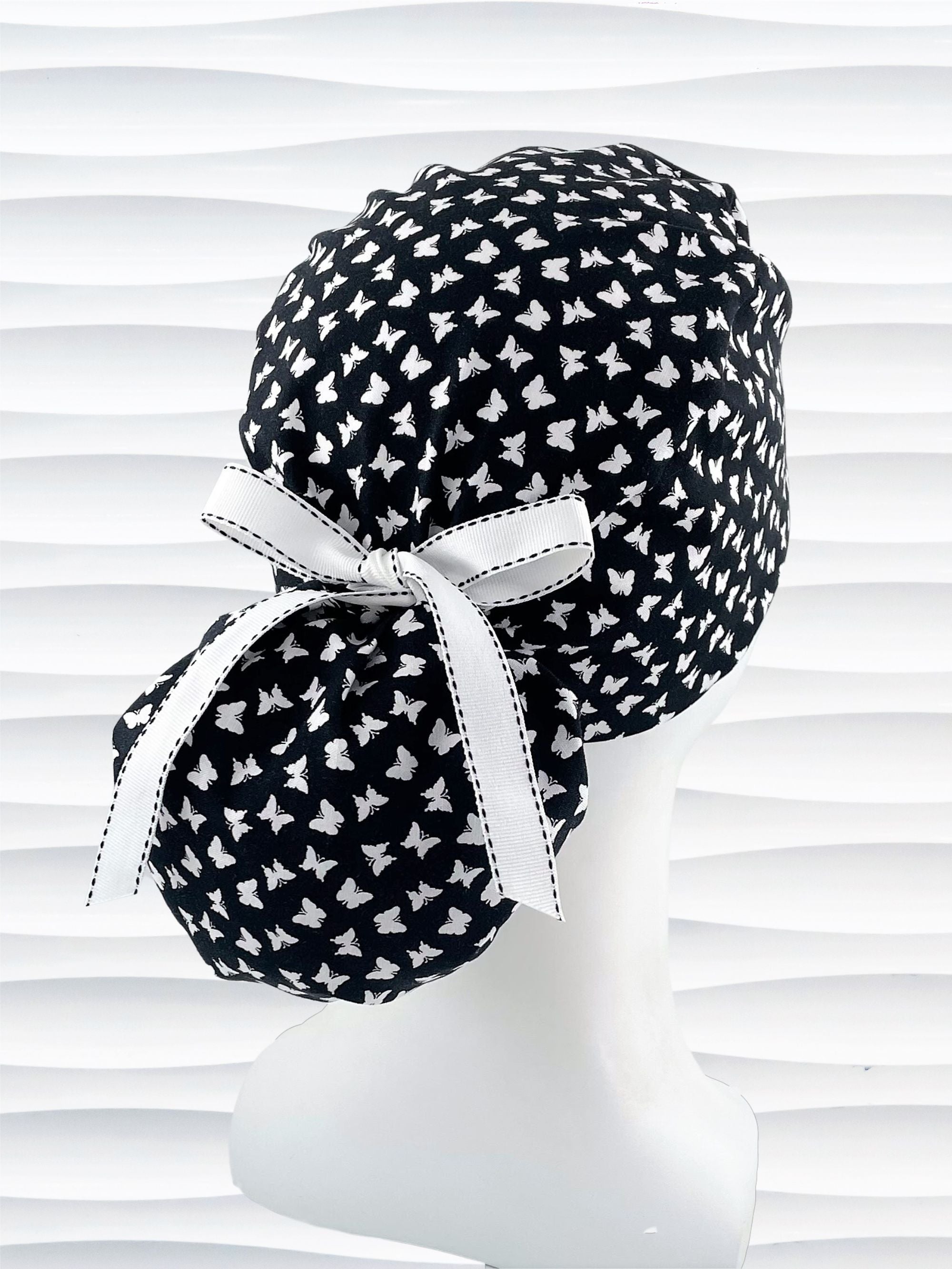 Ponytail style scrub cap hat with white butterflies on black cotton fabric.