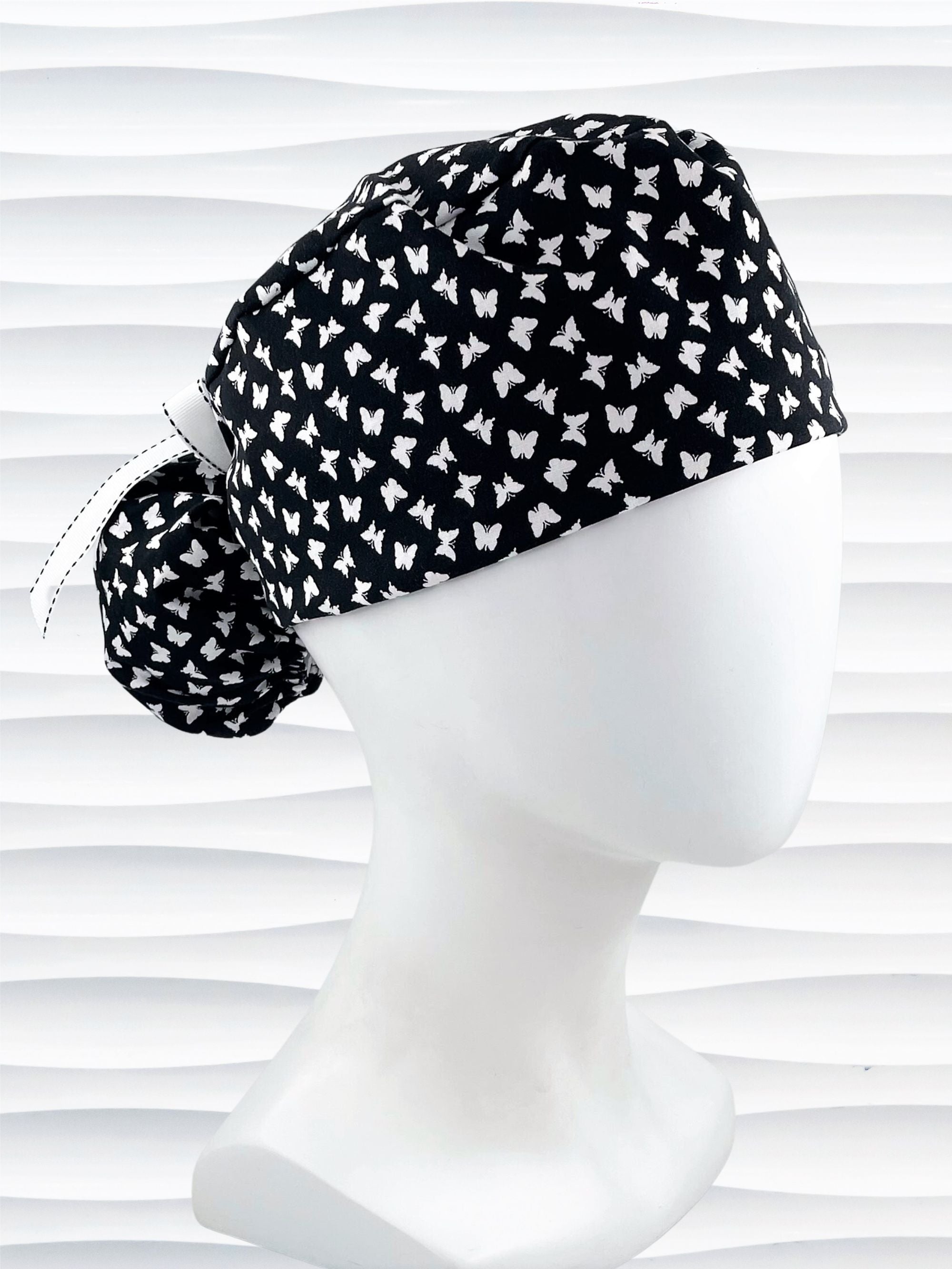 Ponytail style scrub cap hat with white butterflies on black cotton fabric.