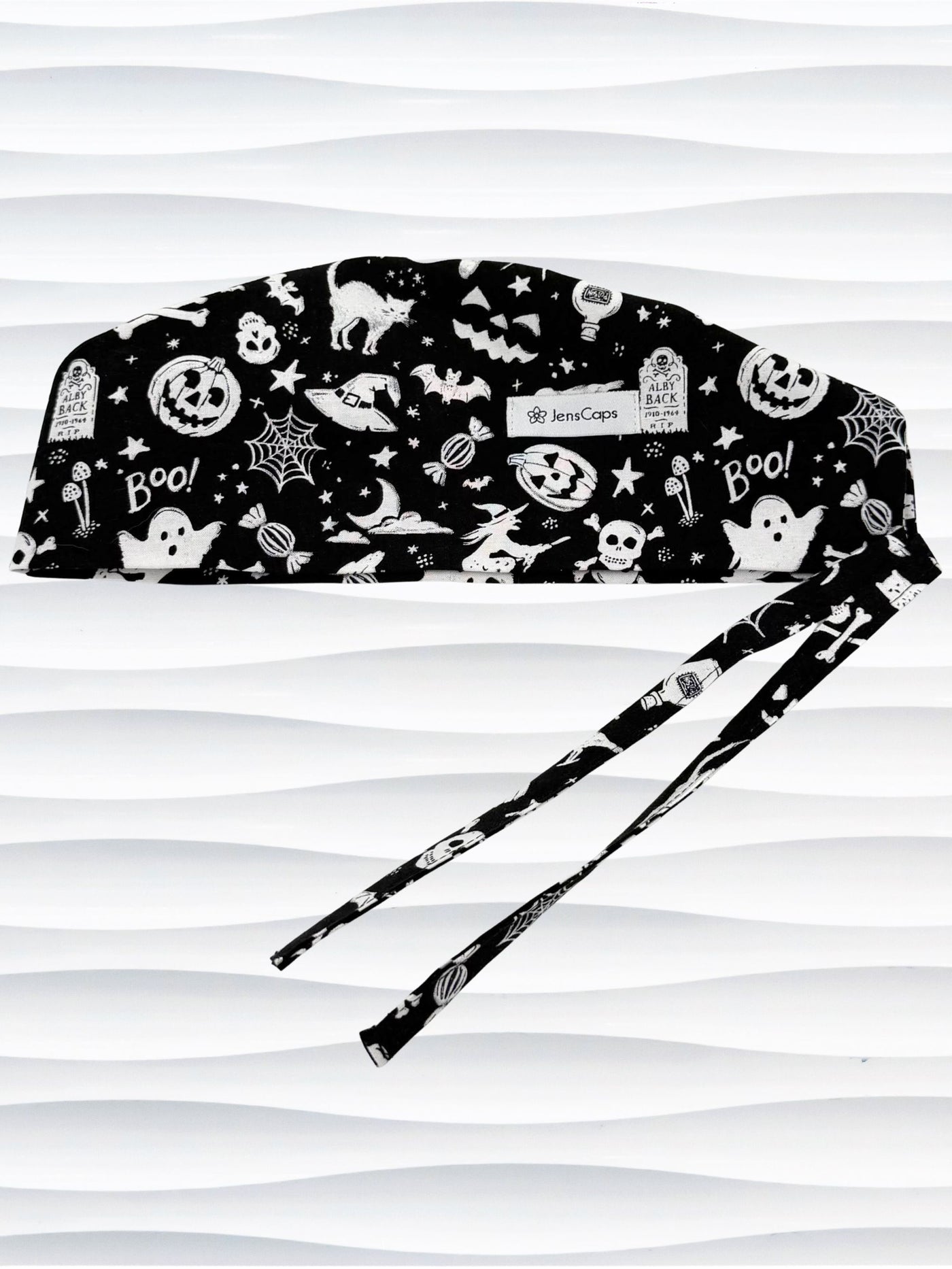 Black and white Halloween-themed scrub cap with a pattern of ghosts, pumpkins, bats, spiders, and stars, featuring adjustable ties, shown against a wavy white background.