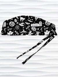 Black and white Halloween-themed scrub cap with a pattern of ghosts, pumpkins, bats, spiders, and stars, featuring adjustable ties, shown against a wavy white background.