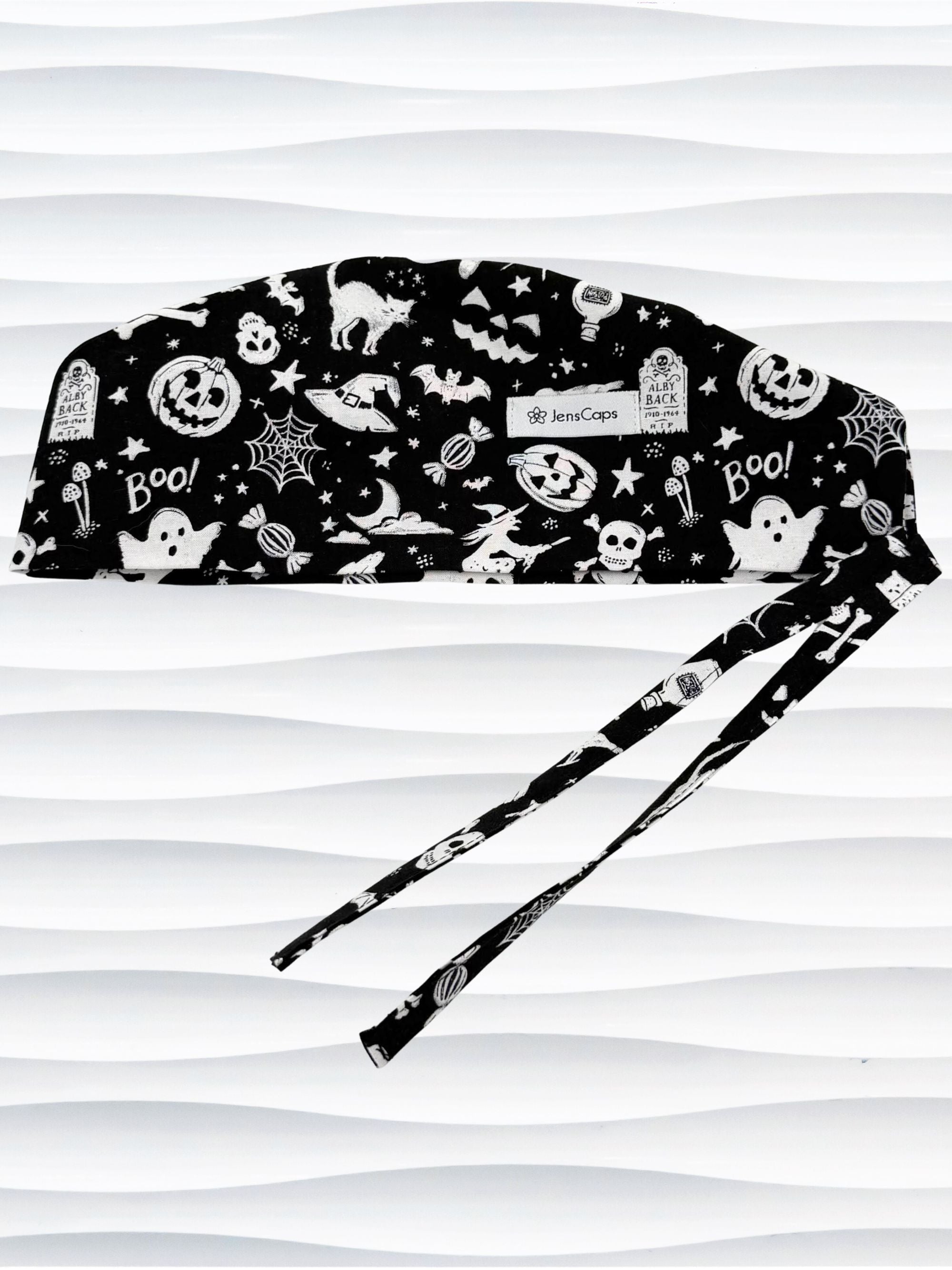 Black and white Halloween-themed scrub cap with a pattern of ghosts, pumpkins, bats, spiders, and stars, featuring adjustable ties, shown against a wavy white background.