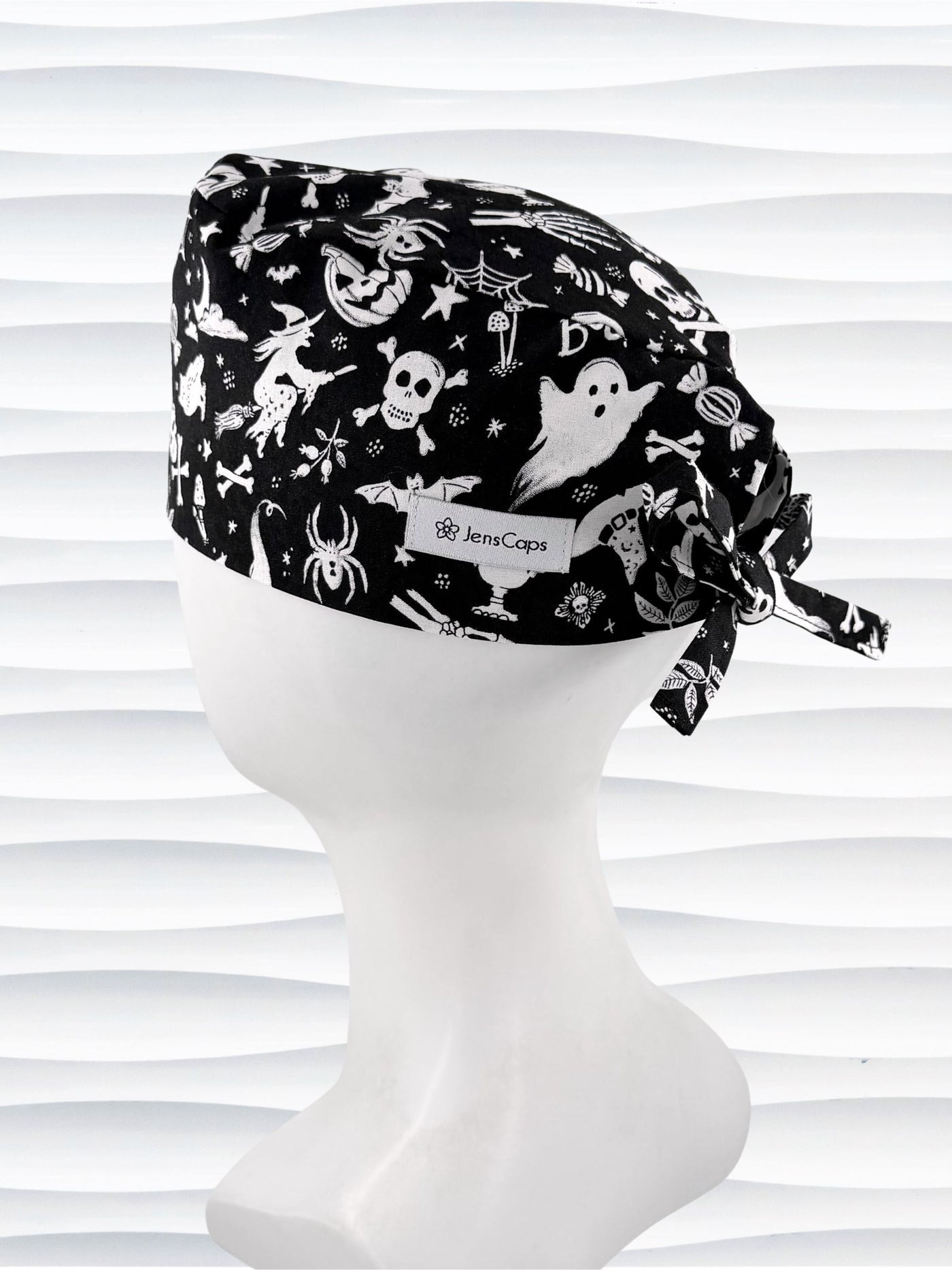 A black tie back style surgical scrub cap by JensCaps with a white Halloween-themed print, displayed on a white mannequin head. The print features ghosts, pumpkins, skulls, and bats.