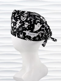 A black tie back style surgical scrub cap by JensCaps with a white Halloween-themed print, displayed on a white mannequin head. The print features ghosts, pumpkins, skulls, and bats.