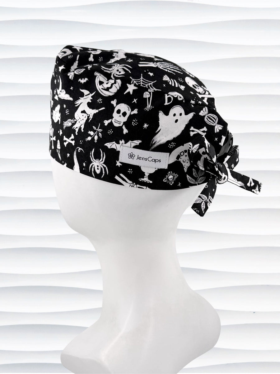 A black tie back style surgical scrub cap by JensCaps with a white Halloween-themed print, displayed on a white mannequin head. The print features ghosts, pumpkins, skulls, and bats.
