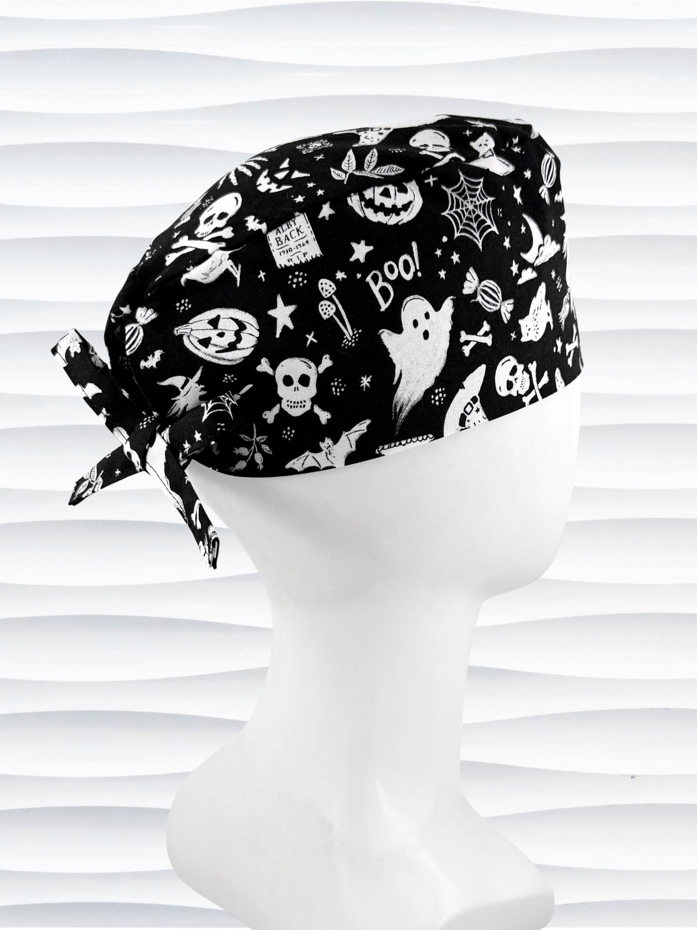 A black tie back style surgical scrub cap by JensCaps with a white Halloween-themed print, displayed on a white mannequin head. The print features ghosts, pumpkins, skulls, and bats.