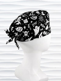 A black tie back style surgical scrub cap by JensCaps with a white Halloween-themed print, displayed on a white mannequin head. The print features ghosts, pumpkins, skulls, and bats.