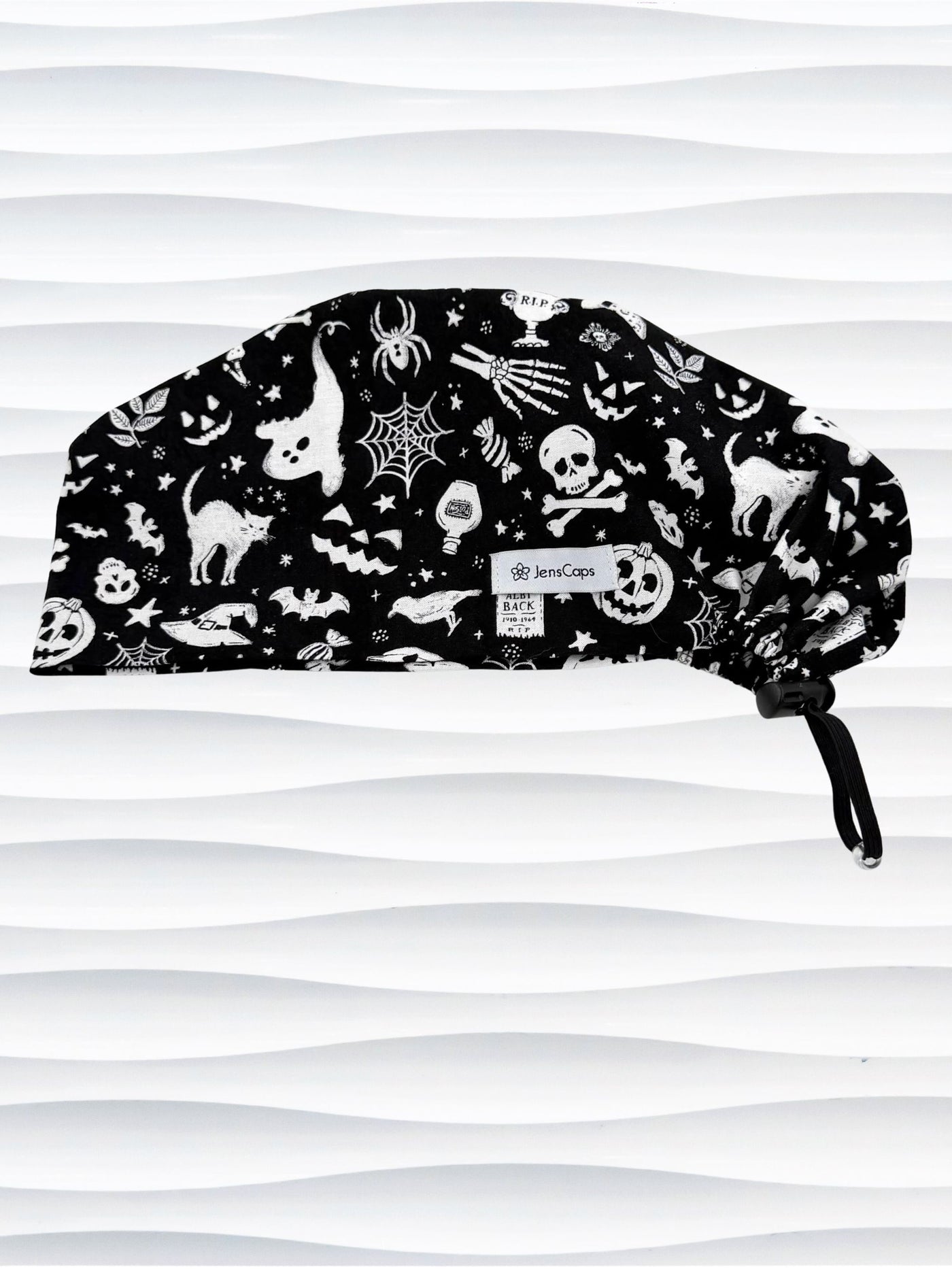 A black surgical scrub cap with a white Halloween-themed print featuring cats, skulls, spiders, and pumpkins, shown against a wavy white background.
