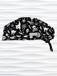 A black surgical scrub cap with a white Halloween-themed print featuring cats, skulls, spiders, and pumpkins, shown against a wavy white background.