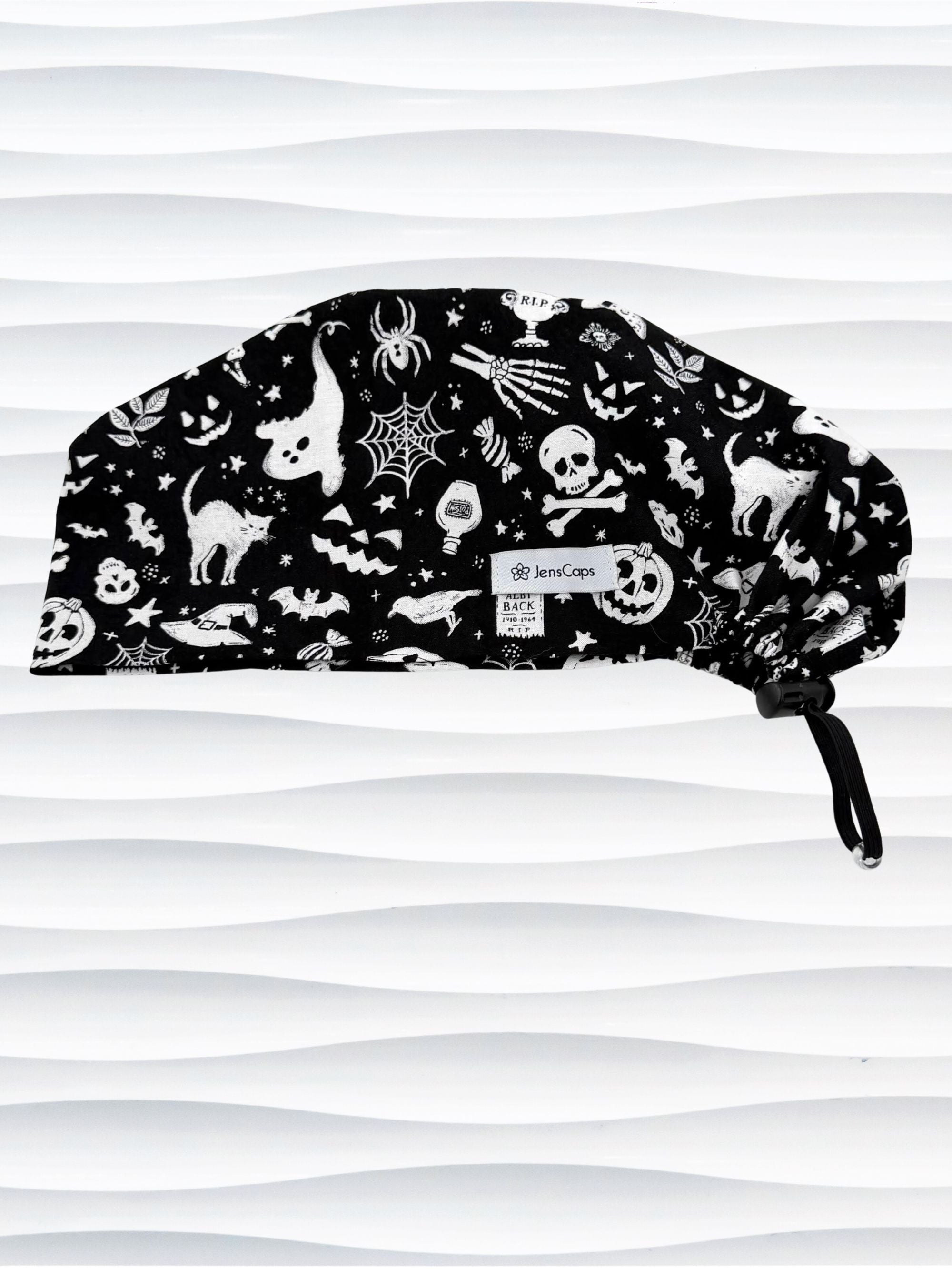 A black surgical scrub cap with a white Halloween-themed print featuring cats, skulls, spiders, and pumpkins, shown against a wavy white background.