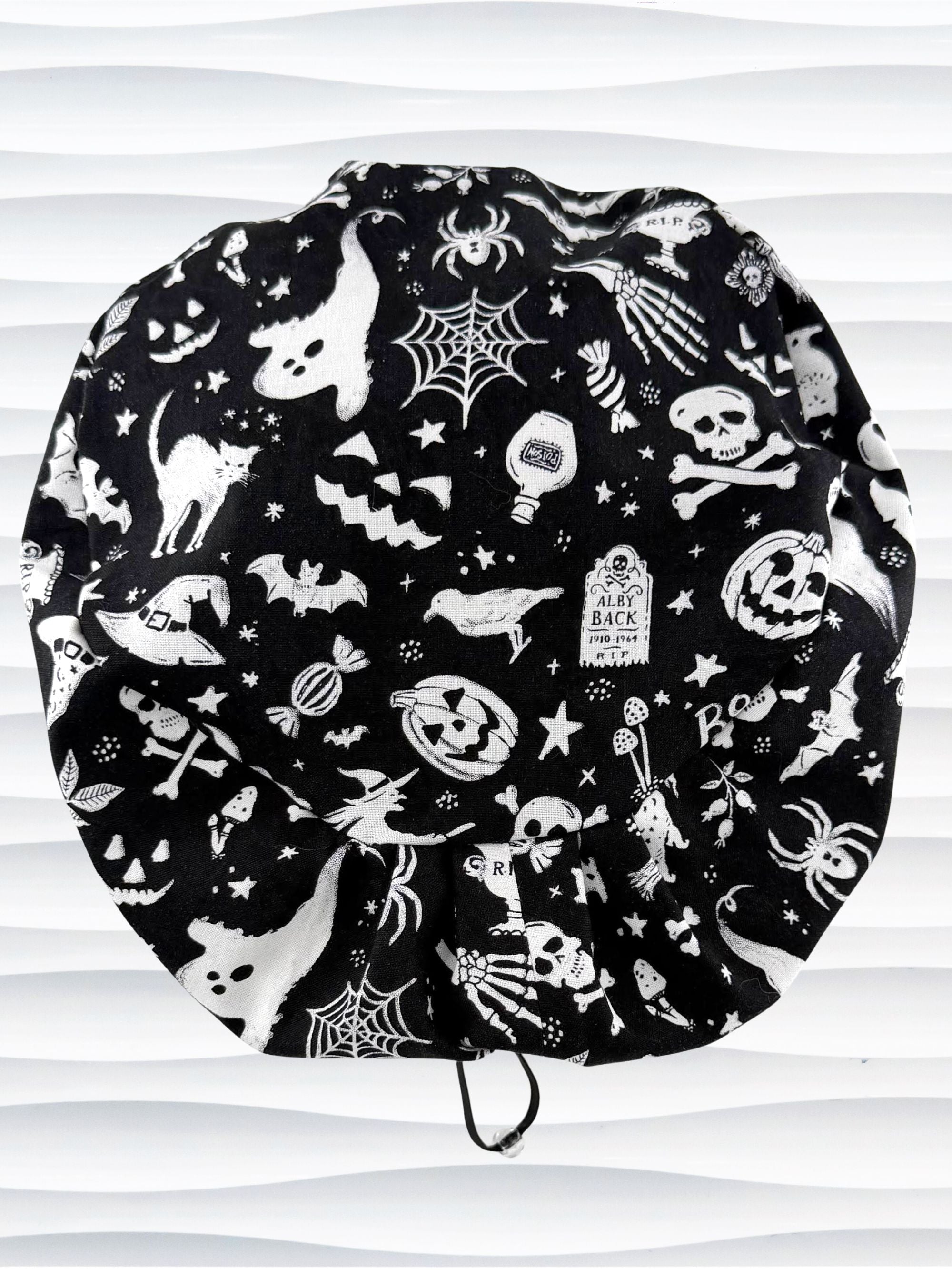 A black fabric Euro Pixie style surgical scrub cap by JensCaps with a white Halloween-themed print of ghosts, bats, spiders, and jack-o'-lanterns, displayed on a white mannequin head 